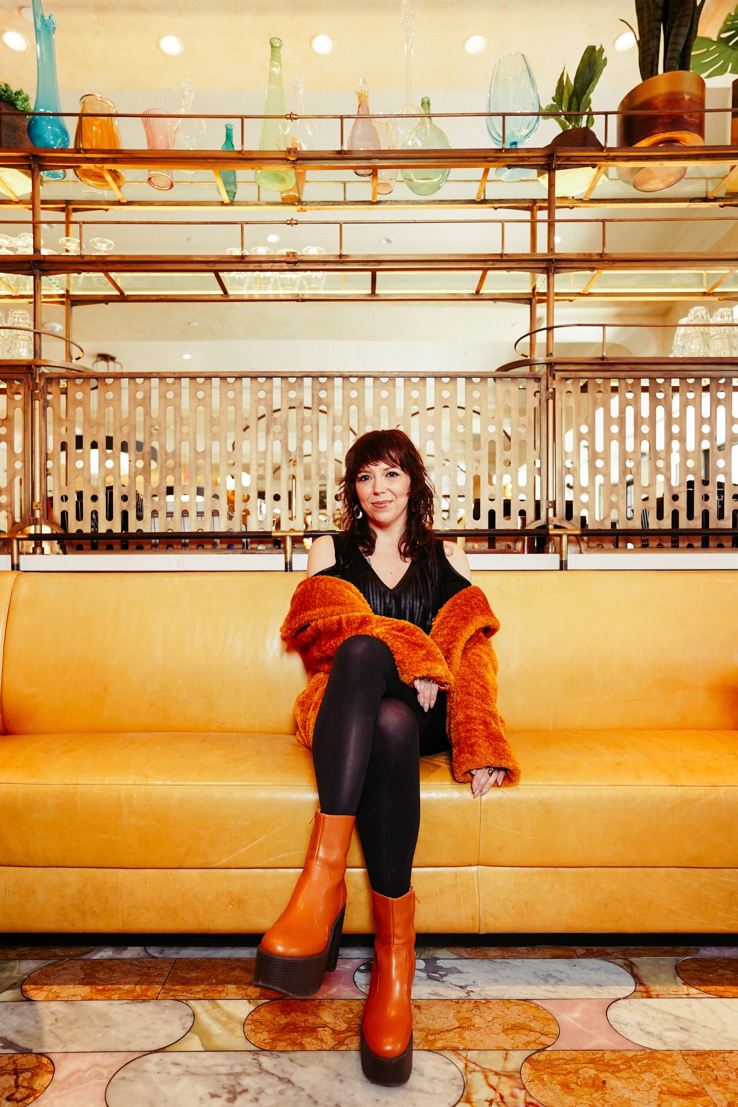Alina Pleskova seated on a cream colored couch in front of pink Art Deco style paneling